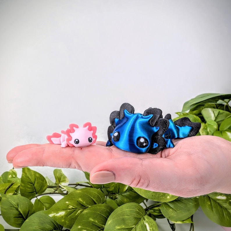 May include: Two small axolotl figurines, one pink and one blue, are sitting on a hand. The pink axolotl has a small, curled tail. The blue axolotl has a larger, straight tail. Both axolotls have large, black eyes.