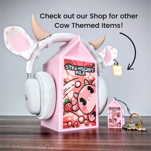 May include: Pink cow-themed headphones with a pink milk carton stand. The milk carton has a cartoon cow and the words "Strawberry Milk" on it. There is a smaller pink milk carton keychain with a cow on it.