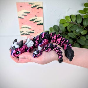 K&ouml;nnte beinhalten: Ein beweglicher Drachenfigur in Pink, Lila und Schwarz. Der Drache hat wei&szlig;e Fl&uuml;gel und H&ouml;rner. Die Figur wird in einer Hand gehalten, mit einem rosa-wei&szlig;en Gem&auml;lde im Hintergrund. Einzigartiges Dekorationsst&uuml;ck.