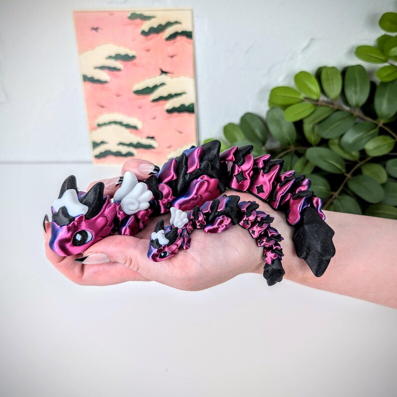 K&ouml;nnte beinhalten: Ein beweglicher Drachenfigur in Pink, Lila und Schwarz. Der Drache hat wei&szlig;e Fl&uuml;gel und H&ouml;rner. Die Figur wird in einer Hand gehalten, mit einem rosa-wei&szlig;en Gem&auml;lde im Hintergrund. Einzigartiges Dekorationsst&uuml;ck.