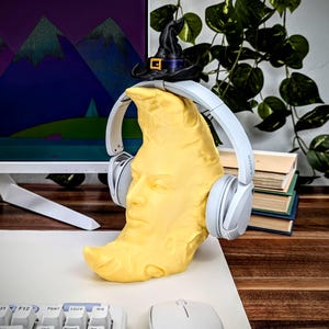 K&ouml;nnte beinhalten: Eine gelbe Halbmondskulptur mit menschlichem Gesicht, die wei&szlig;e Kopfh&ouml;rer und einen Miniatur-Hexenhut tr&auml;gt. Die Kopfh&ouml;rer sind mit "SONY" versehen. Die Skulptur steht auf einer wei&szlig;en Oberfl&auml;che mit Tastatur und Maus im Vordergrund.