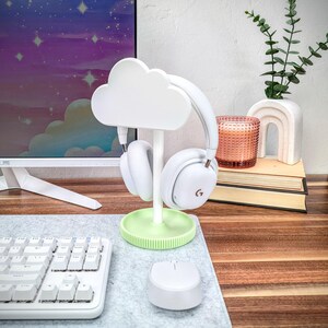 May include: White cloud-shaped headphone stand with a green base. The stand is holding a pair of white headphones. The stand is on a wooden desk with a keyboard and a white mouse.