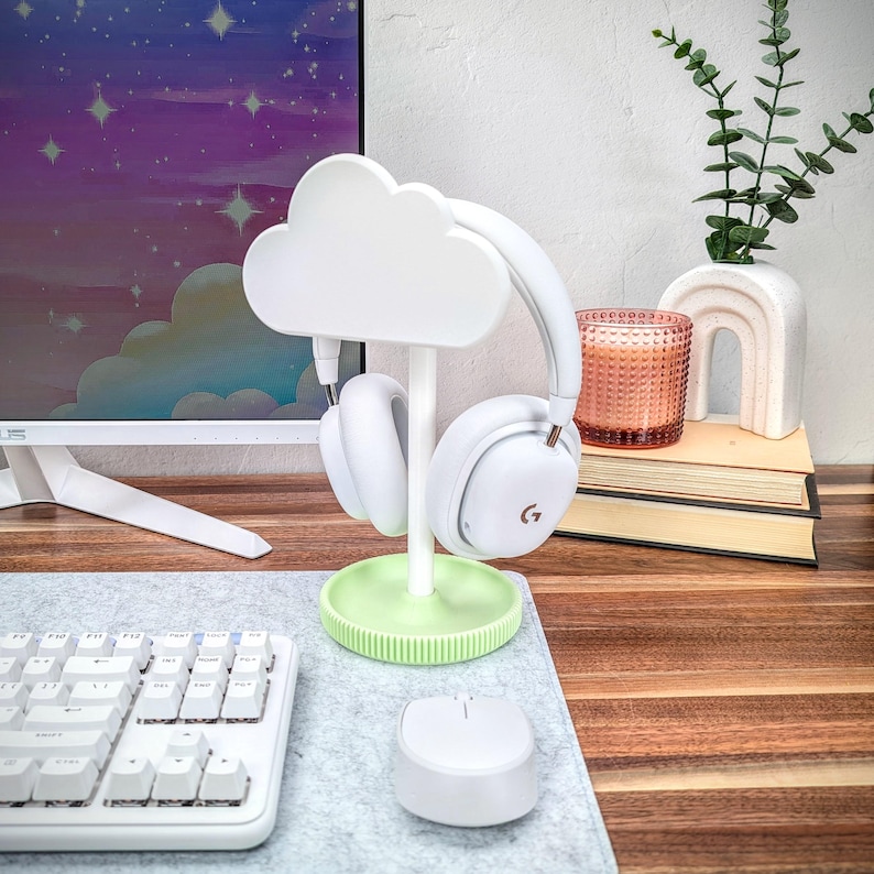 May include: White cloud-shaped headphone stand with a green base. The stand is holding a pair of white headphones. The stand is on a wooden desk with a keyboard and a white mouse.