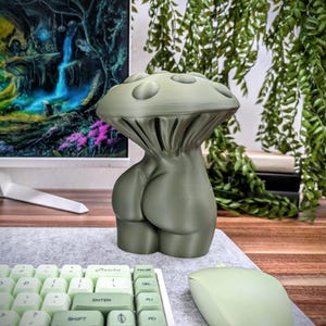 May include: A green 3D printed mushroom-shaped sculpture with a human-like body. The sculpture is sitting on a desk with a keyboard and mouse.