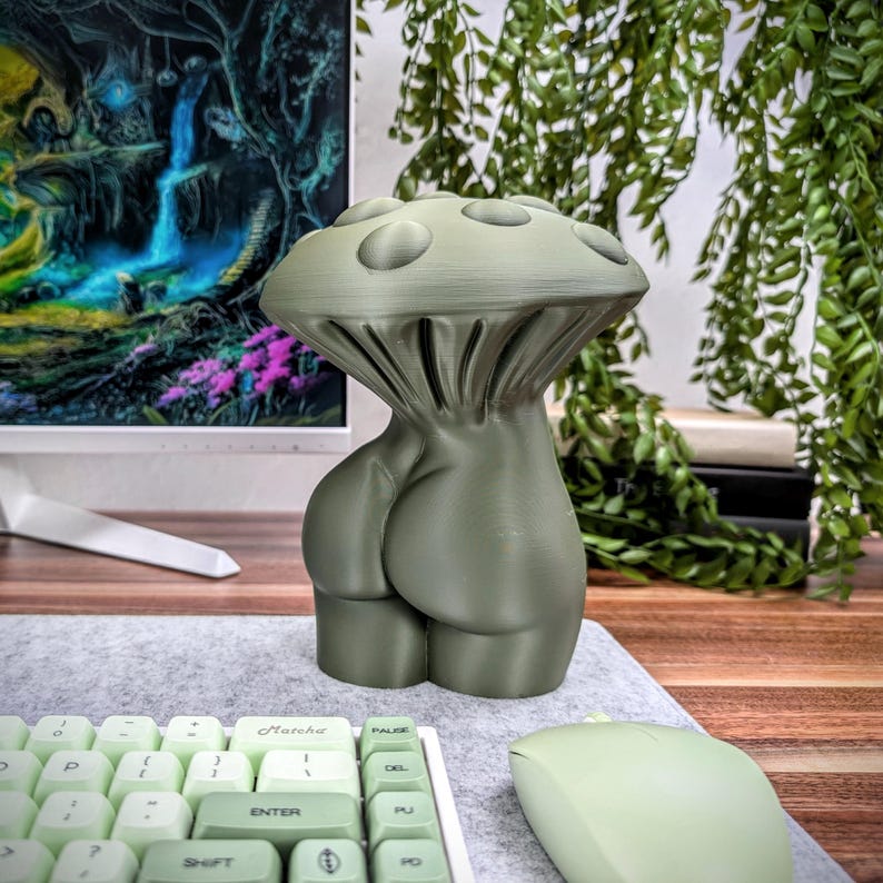May include: A green 3D printed mushroom-shaped sculpture with a human-like body. The sculpture is sitting on a desk with a keyboard and mouse.
