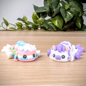 May include: Two small, colorful, hand-painted figurines. One is a rainbow-colored creature with a white fluffy head. The other is a white creature with purple spots and a purple mane. Both have large, black eyes.