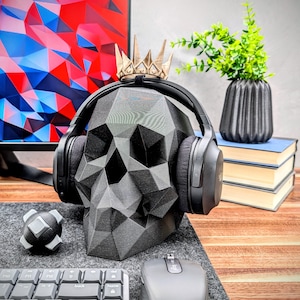 May include: A black skull-shaped headphone stand with a gold crown on top. The stand is holding a pair of black over-ear headphones. The stand is on a wooden desk with a keyboard, mouse, and a small gray object.