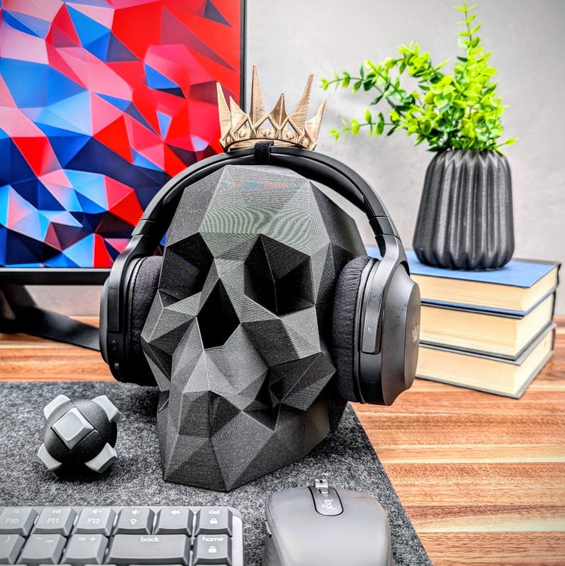 May include: A black skull-shaped headphone stand with a gold crown on top. The stand is holding a pair of black over-ear headphones. The stand is on a wooden desk with a keyboard, mouse, and a small gray object.