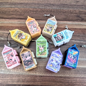 May include: Nine colourful miniature milk carton keychains with different flavours and designs. Each keychain features a cartoon cow and the name of the flavour. The flavours include Strawberry, Chocolate, Matcha Latte, Oat Milk, Peach & Cream, Space Milk, and La Vendor Milk.
