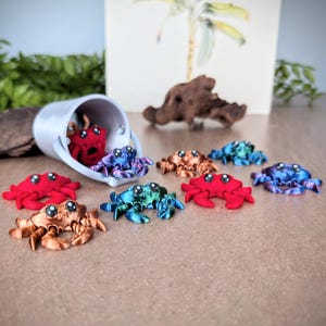 May include: A collection of small, colorful, 3D printed crabs. The crabs are in various shades of red, blue, and purple. Some of the crabs are in a small white bucket, while others are scattered on a brown surface.