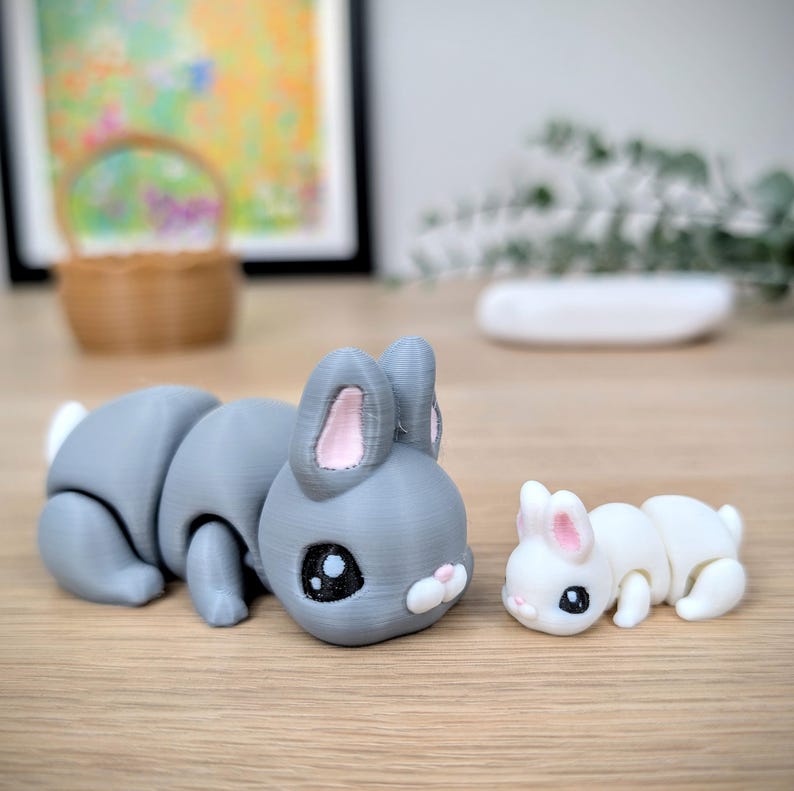 May include: Two articulated bunny figures. One is grey with pink inner ears and a white tail, the other is white with pink inner ears. Both have black eyes and are lying down.