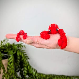 May include: Two red plush lobster toys with white spots. The larger lobster is on a person's hand, while the smaller lobster is on the person's finger.