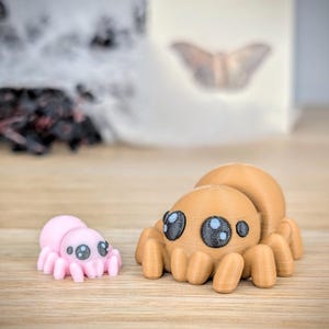 May include: Two 3D-printed spider figurines. One is light pink, the other is tan. Both have large, cartoonish eyes with black pupils and blue highlights. The spiders are posed on a wooden surface.