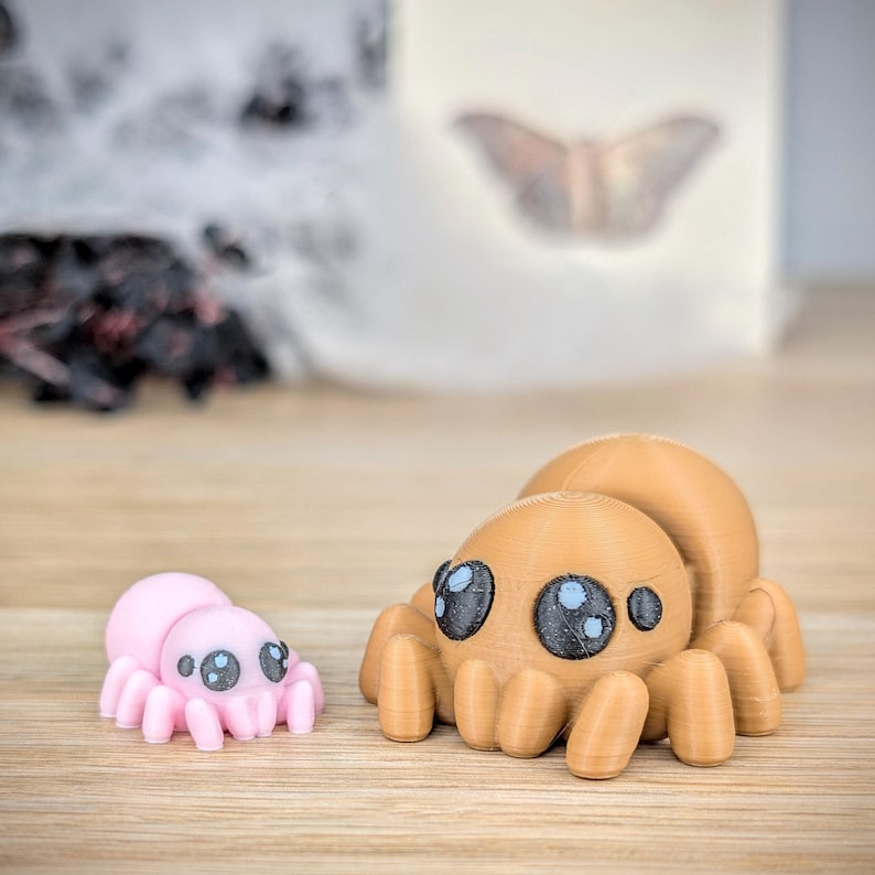 May include: Two 3D-printed spider figurines. One is light pink, the other is tan. Both have large, cartoonish eyes with black pupils and blue highlights. The spiders are posed on a wooden surface.