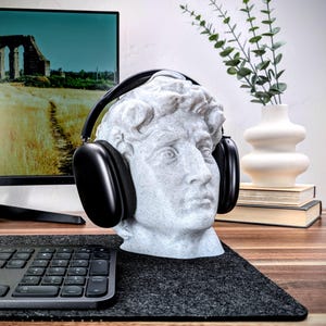 May include: A white plaster bust of a man wearing black over-ear headphones. The bust is sitting on a black desk mat with a black keyboard in the foreground.