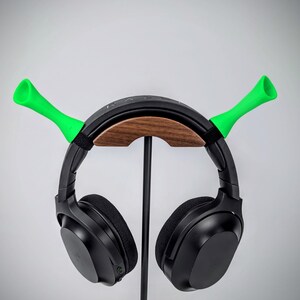 May include: Black headphones with a wooden and black stand, featuring bright green horn-shaped accents. The stand is supported by a black metal pole. The headphones are resting on the stand against a plain white background.