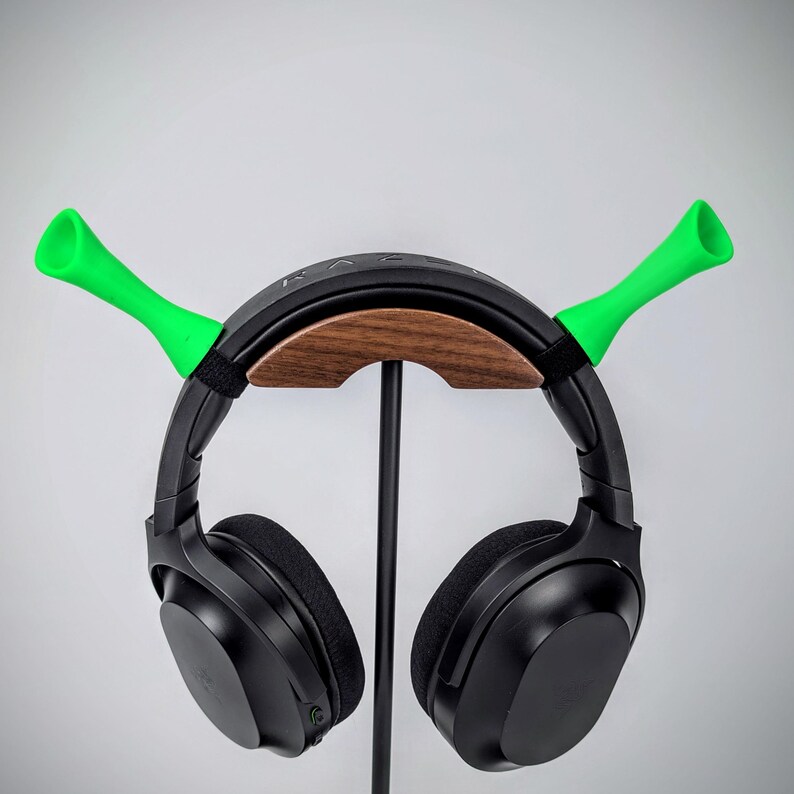 May include: Black headphones with a wooden and black stand, featuring bright green horn-shaped accents. The stand is supported by a black metal pole. The headphones are resting on the stand against a plain white background.