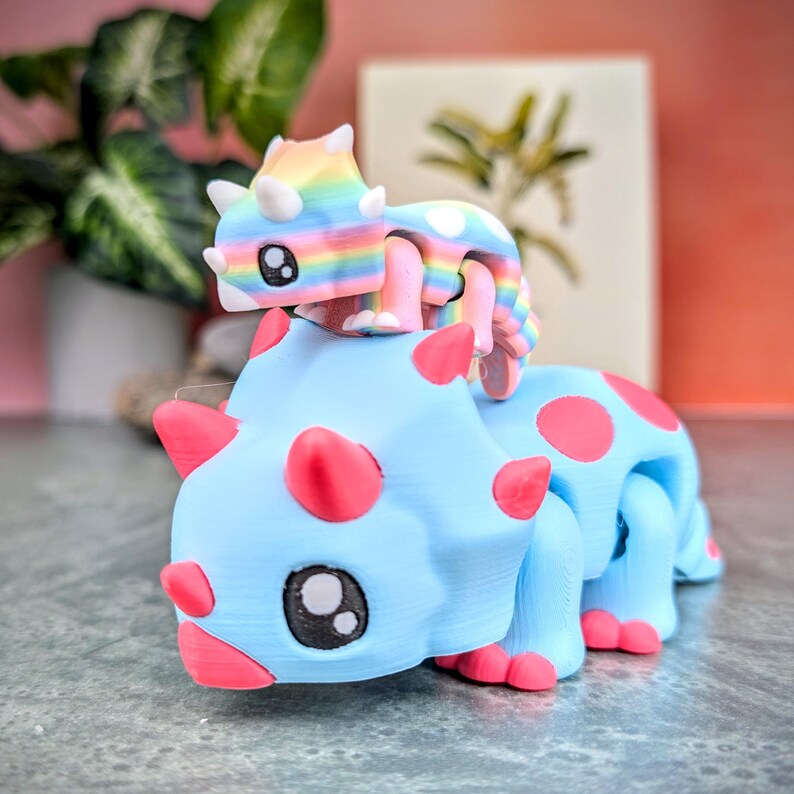 May include: A blue and pink dinosaur toy with a rainbow dinosaur toy riding on its back. Both dinosaurs have large eyes and a friendly expression.