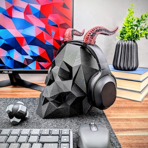 May include: A black skull-shaped headphone stand with red horns. The stand is made of a 3D printed material and has a geometric design. The headphones are black with a silver headband.