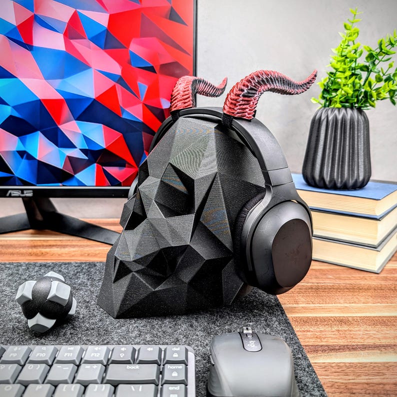May include: A black skull-shaped headphone stand with red horns. The stand is made of a 3D printed material and has a geometric design. The headphones are black with a silver headband.