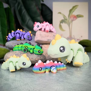 May include: A collection of colorful, articulated dinosaur figurines in various shades of green, purple, pink, and rainbow. Each dinosaur has a unique design with yellow accents and large, black eyes. The figurines are posed on small rocks.