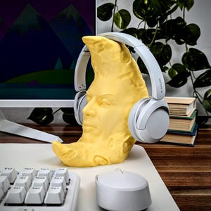 K&ouml;nnte beinhalten: Ein gelber, mondf&ouml;rmiger Kopfh&ouml;rerst&auml;nder mit einem geformten Gesicht, der ein Paar graue Sony-Kopfh&ouml;rer h&auml;lt. Der St&auml;nder steht auf einem wei&szlig;en Schreibtisch neben einer wei&szlig;en Tastatur und Maus. Ein Computermonitor und Gr&uuml;nzeug sind im Hintergrund.