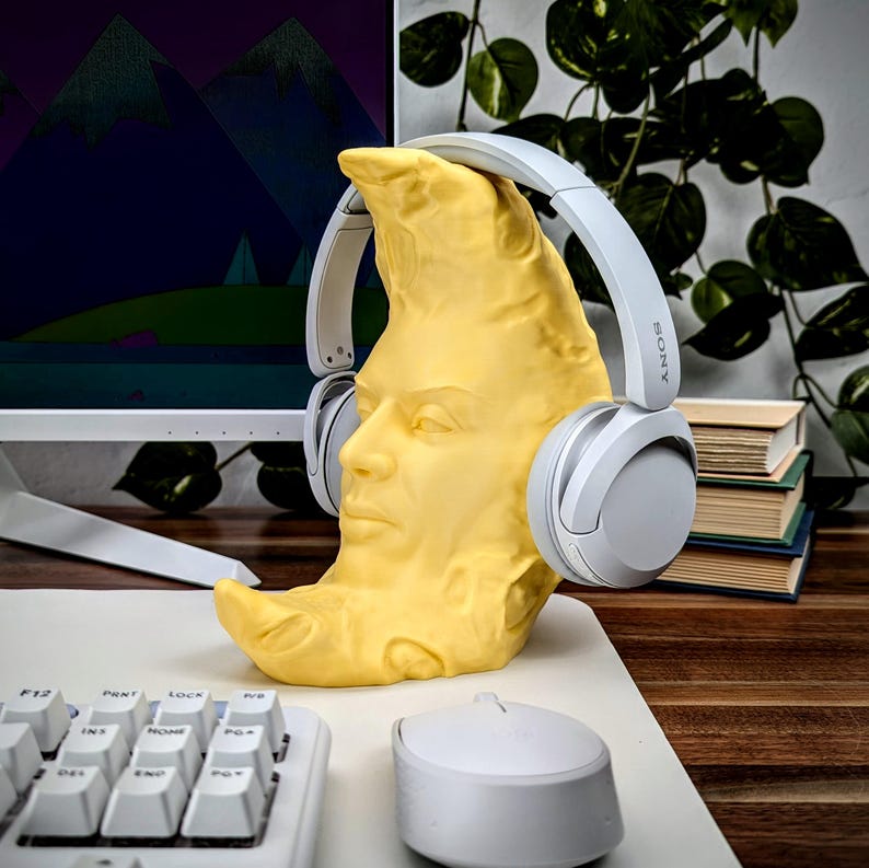 K&ouml;nnte beinhalten: Ein gelber, mondf&ouml;rmiger Kopfh&ouml;rerst&auml;nder mit einem geformten Gesicht, der ein Paar graue Sony-Kopfh&ouml;rer h&auml;lt. Der St&auml;nder steht auf einem wei&szlig;en Schreibtisch neben einer wei&szlig;en Tastatur und Maus. Ein Computermonitor und Gr&uuml;nzeug sind im Hintergrund.