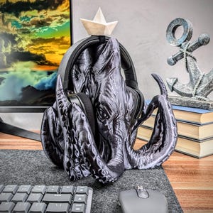 May include: A black 3D printed octopus headphone stand with a small white paper boat on top. The stand is holding a pair of black headphones.