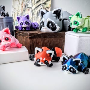May include: A collection of small, hand-painted animal figurines. The figurines are made of a smooth, colorful material and depict various animals, including raccoons, red pandas, and a green creature. The figurines are arranged on a wooden surface and a white surface.