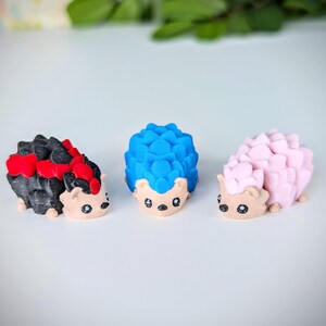 May include: Three small hedgehog figurines, one black with red accents, one blue, and one pink. The hedgehogs have cute faces and are made of a smooth, textured material.