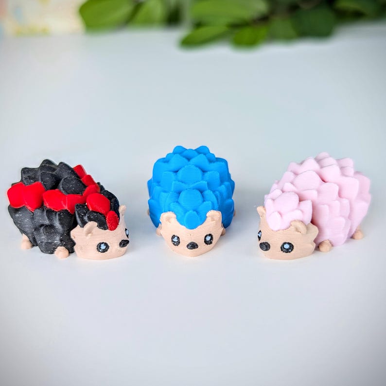 May include: Three small hedgehog figurines, one black with red accents, one blue, and one pink. The hedgehogs have cute faces and are made of a smooth, textured material.