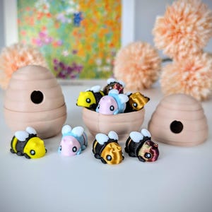 May include: A set of colorful bee figurines, some in a beehive-shaped container, others on a white surface. The bees are made of plastic and have different colors and patterns.