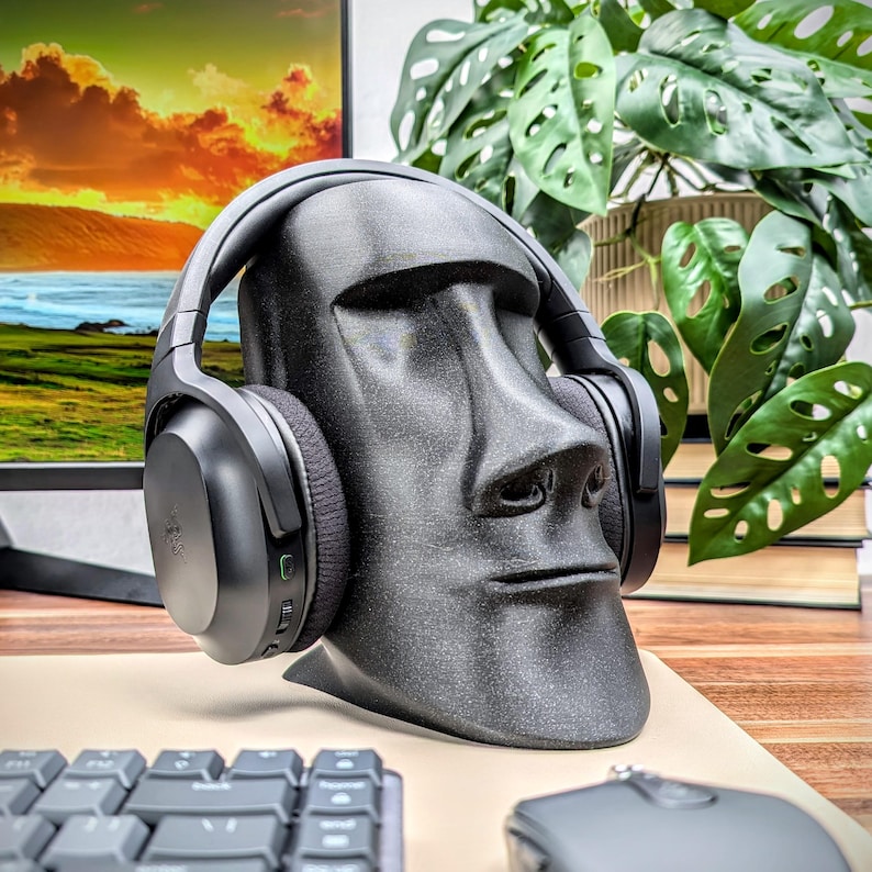 May include: Black over-ear headphones with a microphone are displayed on a black and gray 3D printed stand shaped like a human head. The stand is sitting on a wooden surface with a keyboard and mouse in the background.