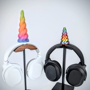 May include: Two white headphones with a unicorn horn on top of each. The unicorn horns are rainbow colored and are made of plastic. The headphones are on a black stand.