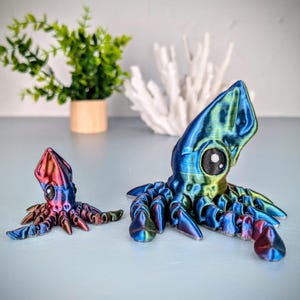 May include: Two iridescent blue and green 3D printed squid figurines. The larger squid has a green head and the smaller squid has a red head. Both squid have black eyes and are posed with their tentacles outstretched.