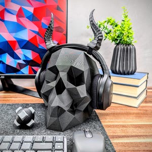 May include: A black skull-shaped headphone stand with horns. The stand is made of a textured, geometric design. The headphones are black with a black headband.