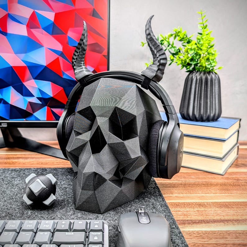 May include: A black skull-shaped headphone stand with horns. The stand is made of a textured, geometric design. The headphones are black with a black headband.