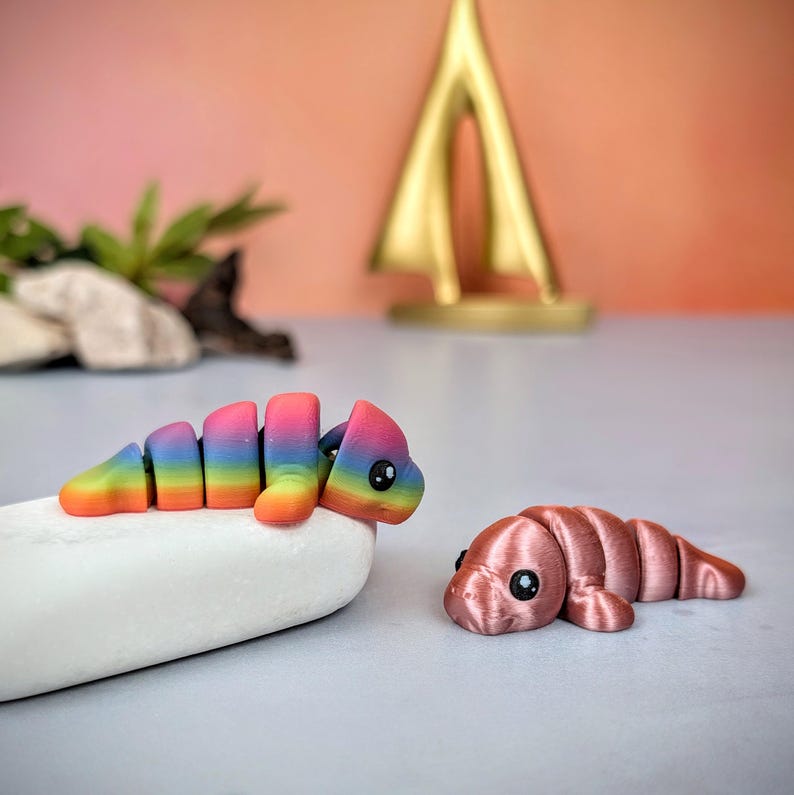May include: Two segmented worm-shaped toys. One is rainbow-coloured, resting on a white stone. The other is a solid pink colour. Both have large, black eyes and a smiling expression. The background is a soft peach colour.