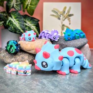 May include: A collection of colorful, 3D printed dinosaur toys. The dinosaurs are posed in various positions and have different colors and patterns. The dinosaurs are sitting on rocks.