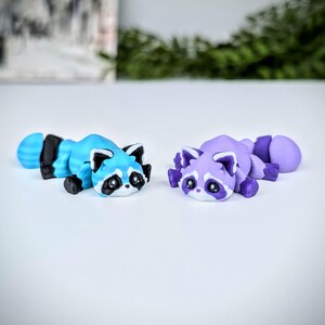 May include: Two small, hand-painted raccoon figurines. One is blue with purple stripes and the other is purple with white stripes.