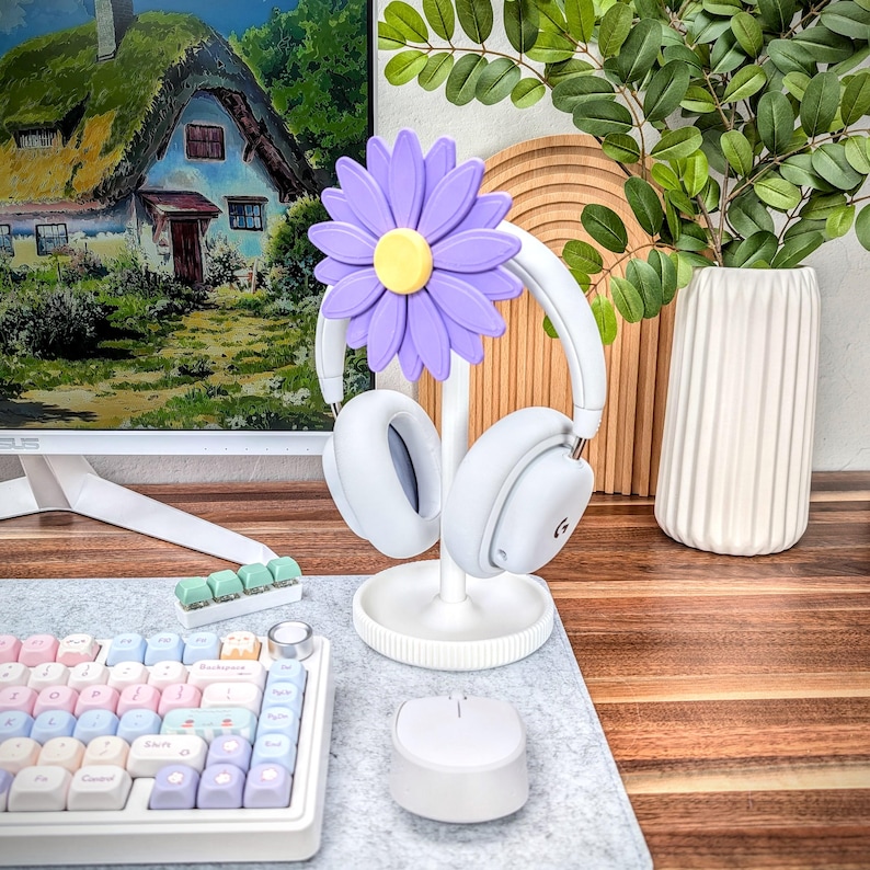 May include: A white headphone stand with a purple flower design. The stand has a round base and a single post that holds the headphones. The headphones are white and have a black microphone.