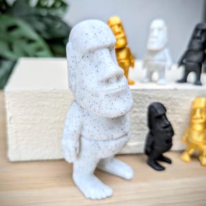 May include: A white speckled figurine of a person with a large head and a simple body. The figurine is standing on a wooden surface.