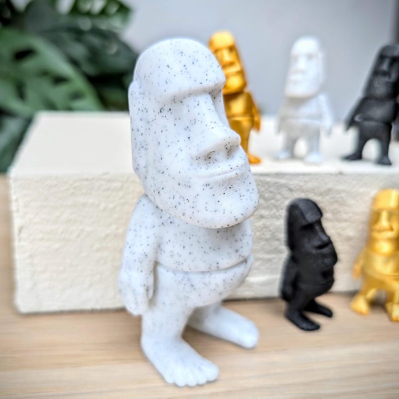 May include: A white speckled figurine of a person with a large head and a simple body. The figurine is standing on a wooden surface.