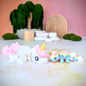 May include: Two small unicorn figurines, one white with pink and gold accents, the other rainbow colored, on a white surface.