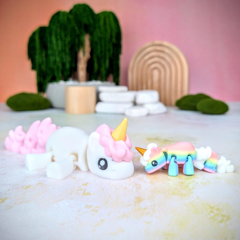 May include: Two small unicorn figurines, one white with pink and gold accents, the other rainbow colored, on a white surface.