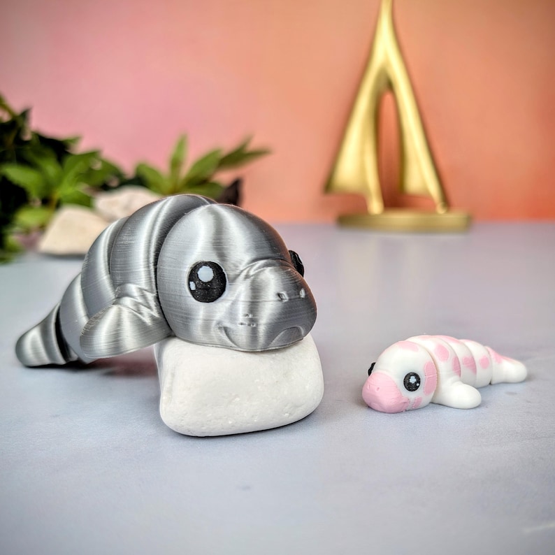 May include: A silver 3D-printed manatee figurine on a white stone, with a smaller pink and white manatee figurine. The larger manatee has large black eyes. A gold abstract sculpture is in the background. The figurines are likely decorative.