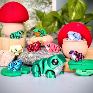 May include: A collection of colorful, miniature frog figurines in various poses. The frogs are in shades of green, red, pink, blue, and purple. Some are perched on rocks, a wooden block, and a lily pad. A red mushroom and a small pot are also in the scene.