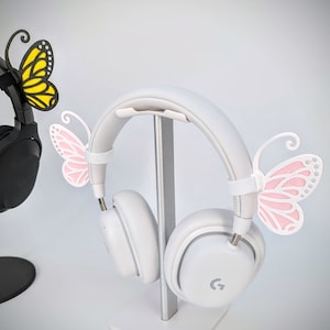 May include: White wireless headphones with a stand and butterfly accents. The headphones have a pink butterfly on one side and a yellow butterfly on the other.
