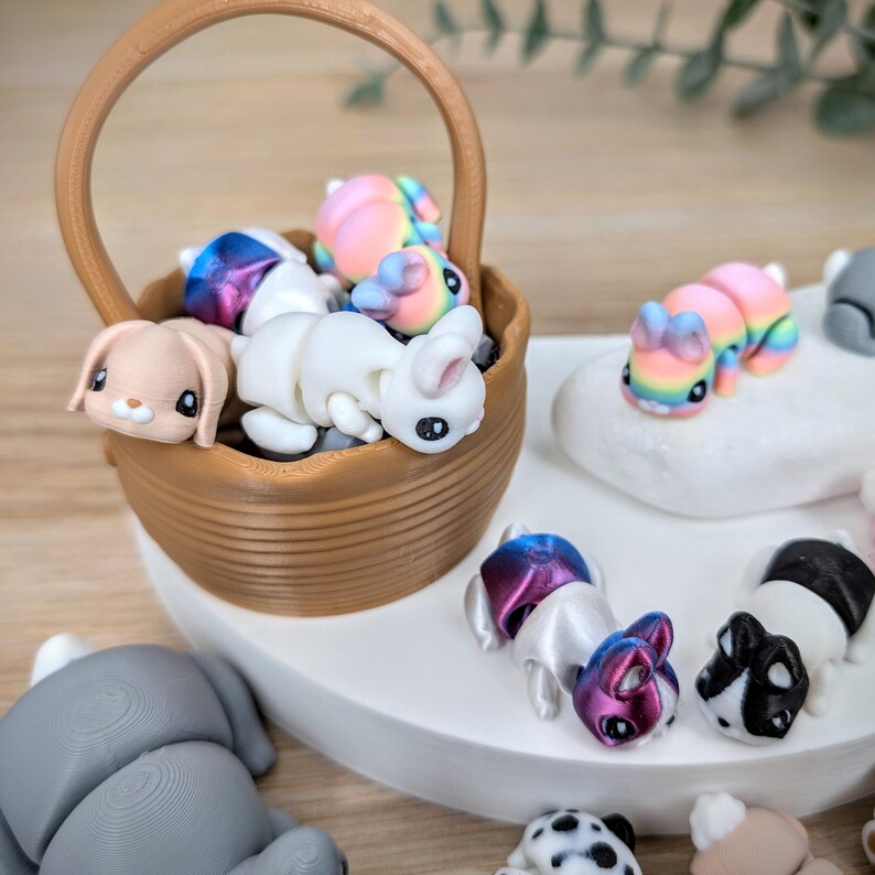 May include: A selection of miniature bunny figurines in various colours. Some are in a brown basket, others are on a white surface. The bunnies are white, pink, blue, and purple, with detailed facial features. The basket is made of a brown material.