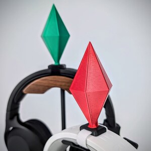 May include: Two 3D printed diamond-shaped objects, one green and one red, are attached to a black and white headset stand. The stand has a wooden base.